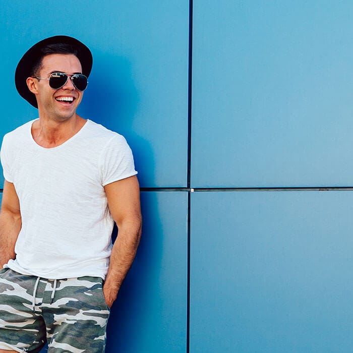 Smiling man in sunglasses and casual clothes against blue wall.
